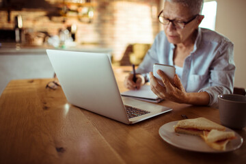 Elderly woman holding smartphone writing notes with laptop at home