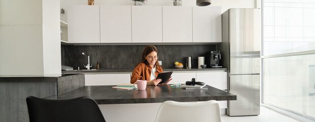 Portrait of young woman, student sitting in kitchen with tablet and notebook, reading news online,...