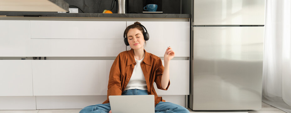 Portrait of young woman, dj mixing sounds on laptop, closes eyes in headphones and listens to music, sits on floor at home