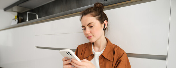 Portrait of young modern woman in wireless headphones, sitting on kitchen floor, using smartphone, making playlist with favourite songs, listens to music
