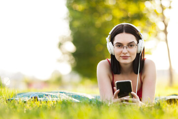 Beautiful dark hair girl listening to music with a headphones from smartphone. Lying on a green meadow. Looking at camera.