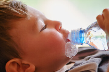 A child drinks water from a blue plastic bottle on the background of a window. Toddler baby with a...