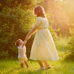 A woman mother dances with a baby holding hands in nature. Happy child with mom on a walk in the summer park. Kid aged about two years (one year eleven months)
