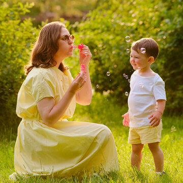 A mother woman plays with a baby in nature blowing soap bubbles. Happy child and mom play on the green grass in the summer park. Kid aged about two years (one year eleven months)