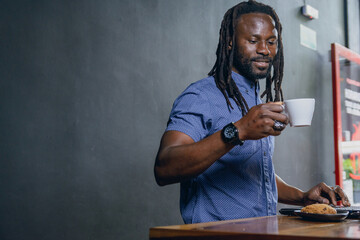 black man of african ethnicity standing working with his laptop inside restaurant and having cup of coffee