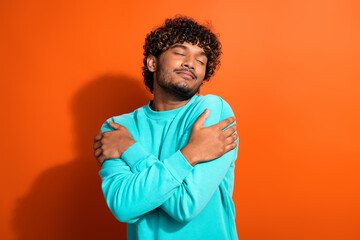 Portrait of young dreaming man curly hair wearing fresh laundry clothing embracing himself shoulders isolated on orange color background