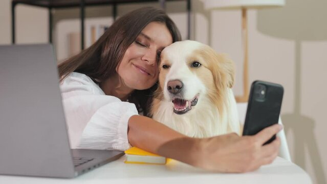 joyful smiling attractive young beautiful freelancer girl takes a selfie and hugging with her cute dog. At home at a desk and computer. Calls via video link with the dog.
