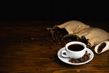 Hot coffee in a white coffee cup and many coffee beans placed around on a wooden table in a warm, light atmosphere, on dark background, with copy space.
