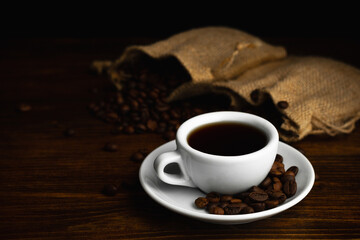 Hot coffee in a white coffee cup and many coffee beans placed around on a wooden table in a warm, light atmosphere, on dark background, with copy space.