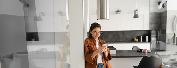 Portrait of cute young woman standing at home with smartphone, reading message on mobile phone,...