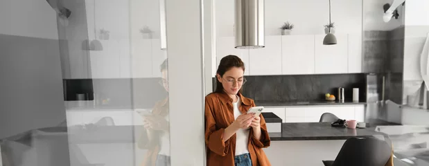 Foto op Canvas Portrait of cute young woman standing at home with smartphone, reading message on mobile phone, using application, order online, paying utility bill in app © Mix and Match Studio