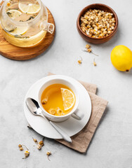 Chamomile herbal tea with lemon in a white cup and teapot with flowers on a light background. The concept of a healthy detox drink for health and sleep.