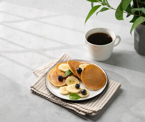 Banana pancakes with fresh fruit slices, berries, mint and honey on a light background with a cup of black coffee, shadows and green branch. Healthy diet breakfast.