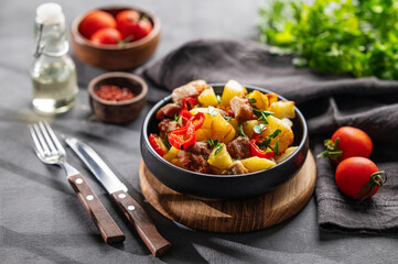 Fried pork meat with potatoes, onions and peppers. National homemade Georgian dish ojakhuri in a black frying pan on a dark background with fresh herbs, spices and tomatoes