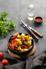 Fried pork meat with potatoes, onions and peppers. National homemade Georgian dish ojakhuri in a black frying pan on a dark background with fresh tomatos, herb and shadows.