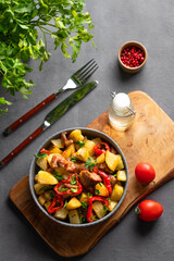 Fried pork meat with potatoes, onions and peppers. National homemade Georgian dish ojakhuri in a bowl on a wooden board on a dark background with spices, fresh tomato and herb.
