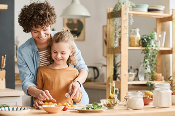 Happy young mom teaching her son to cut vegetables for salad while they cooking together in kitchen
