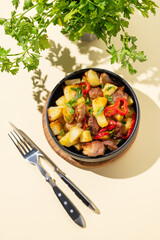 Fried pork meat with potatoes, onions and peppers. National homemade Georgian dish ojakhuri in a black bowl on a yellow background with fresh herb and shadows.
