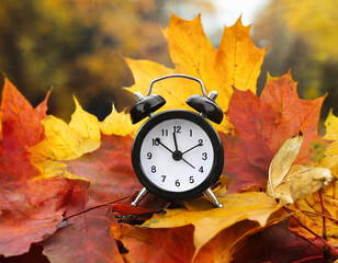 Black alarm clock on colorful maple leaves background. Closeup. Time change concept