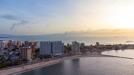 Fototapeta premium Drone photos of the City of Lechería on a sunset, in which you can see buildings, sea, people bathing on the beach, and passers-by. Playa los Canales Boulevard