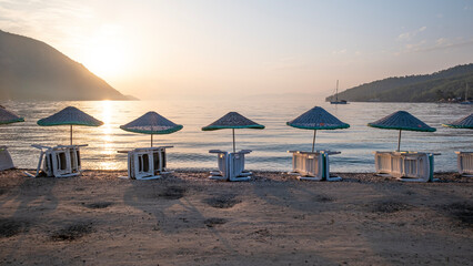 Empty wooden sun loungers and umbrellas on the beach, behind them the mountain covered with green forests, no people, Clean and clear Aegean sea Muğla - Turkey