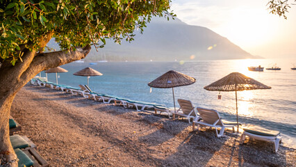 Empty wooden sun loungers and umbrellas on the beach, behind them the mountain covered with green...