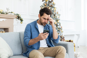 Fototapeta premium Upset unhappy man sitting near Christmas tree on winter day, got bad news on phone, celebrating new year and Christmas, unhappy reading, using app on smartphone, living room at home on sofa