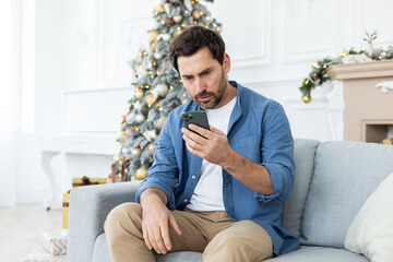 Upset unhappy man sitting near Christmas tree on winter day, got bad news on phone, celebrating new...