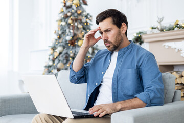 Frustrated sad and unhappy man sitting on sofa in living room near Christmas tree, businessman working remotely on New Year holidays, looking at laptop confused