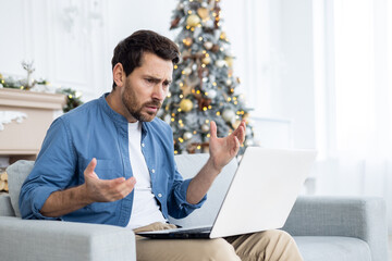 Frustrated sad and unhappy man sitting on sofa in living room near Christmas tree, businessman working remotely on New Year holidays, looking at laptop confused - obrazy, fototapety, plakaty