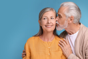 Senior man kissing his beloved woman on light blue background, space for text