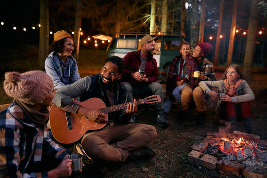 Happy black man plays acoustic guitar while camping with his family and friends in nature at night.