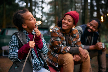 Black little girl eating roasted marshmallows while camping with parents in woods.