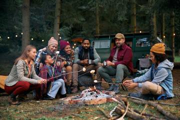 Multiracial families roasting marshmallows while gathering around campfire in woods.
