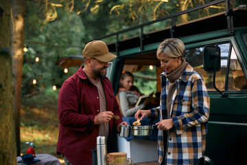 Happy parents cooking during family camping trip in woods. - obrazy, fototapety, plakaty