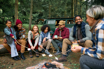 Two happy families relaxing by campfire and roasting marshmallows while camping in nature. - obrazy, fototapety, plakaty