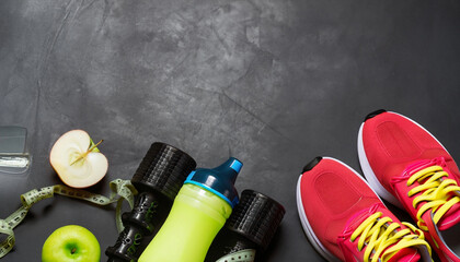 Fitness concept with red and yellow sneakers dumbbells pomelo bottle of water apple and measure tape on black concrete background