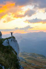 A man standing on the edge of a mountain cliff with arms outstretched at sunset.