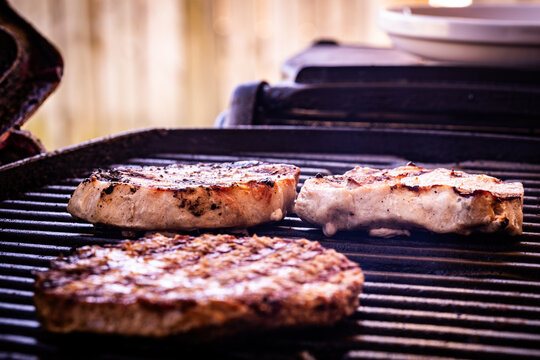 Two Pork Chops Positioned On A Barbecue For Grilling, Placed Behind A Beef Burger Patty With Grill Marks On One Side.