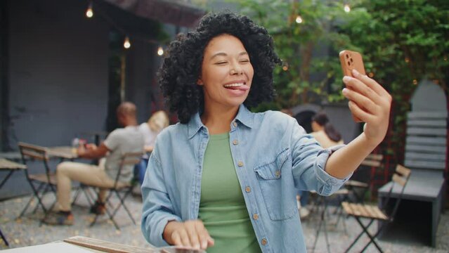 Happy African American woman making selfie on her smartphone while sitting at table with laptop. Pretty curly girl posing on phone camera and cheerfully smiling during photo session in cafe.
