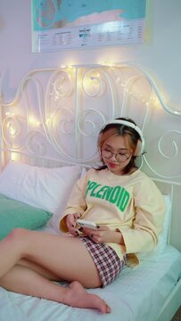 Smiling young asian woman in eyeglasses listening music in headphones while texting on mobile on the bed at home