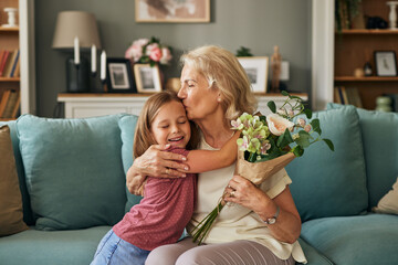 Happy grandma hugging and kissing granddaughter thanking for present, holding flower bouquet