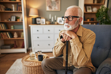 Old disabled man holding hands on wooden stick, resting in cozy armchair in living room