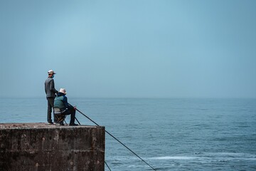 Two fishermen sitting on a pier in front of the water on a gloomy day