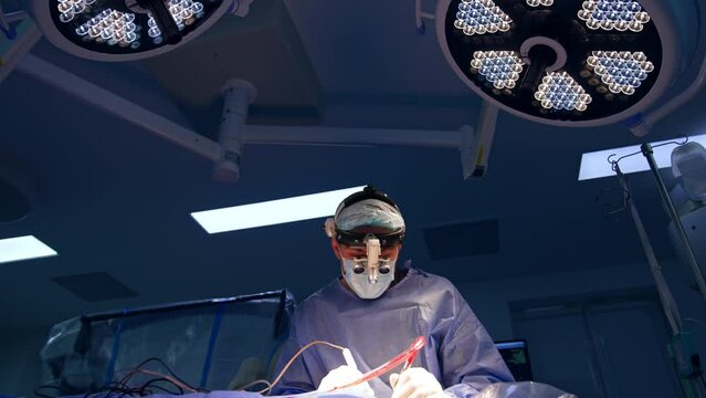 Focused male surgeon wearing device glasses operating the patient. Doctor uses tools holding them in both hands. Low angle view.