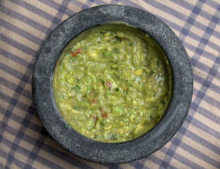 guacamole inside molcajete (traditional mexican mortar and pestle for grinding spices and making sauces) spicy avocado dip with aztec roots on striped tablecloth restaurant table (checkered, boxed)