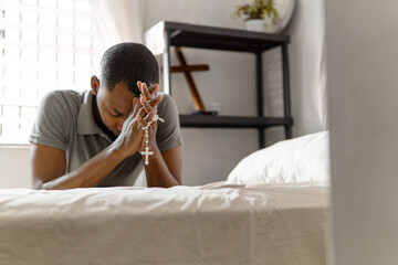 Man holding and praying rosary over a bed at home interior. Christian life concept.
