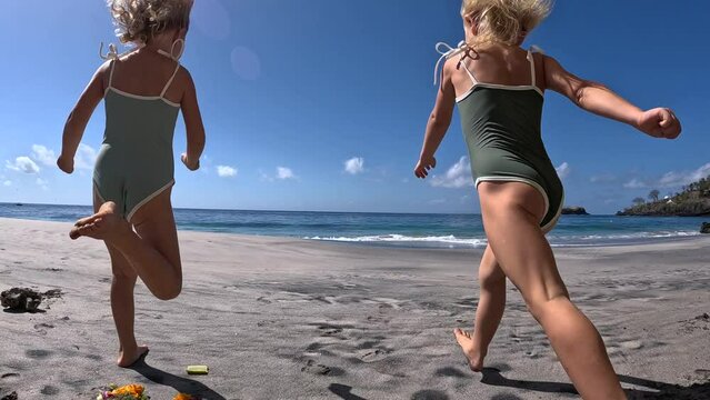 Bottom view of two running happy girls in swimsuits going to swim in ocean. Dynamic video of cute kids running on warm sand past sun loungers and sun umbrellas to swim. Concept of summer swim in ocean