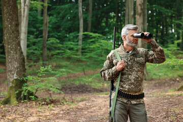 Man with hunting rifle looking through binoculars in forest. Space for text