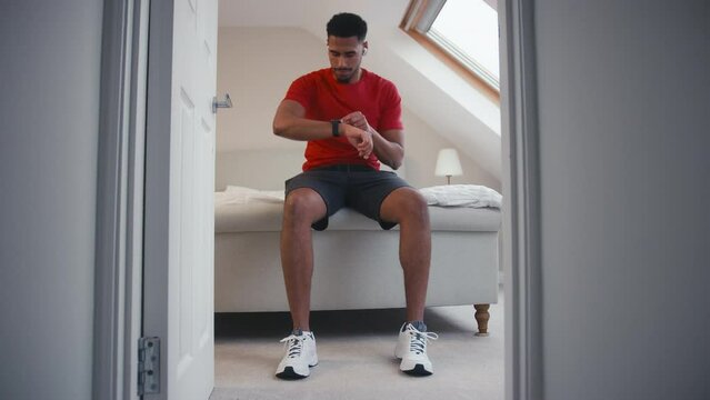 Man in fitness clothing sitting on bed at home wearing wireless ear buds checking fitness app on smart watch before exercise - shot in slow motion